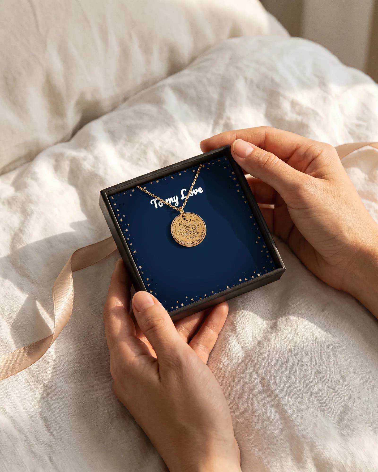 Hands holding the open gift box on a bed: the star map necklace on a navy card that reads To my Love