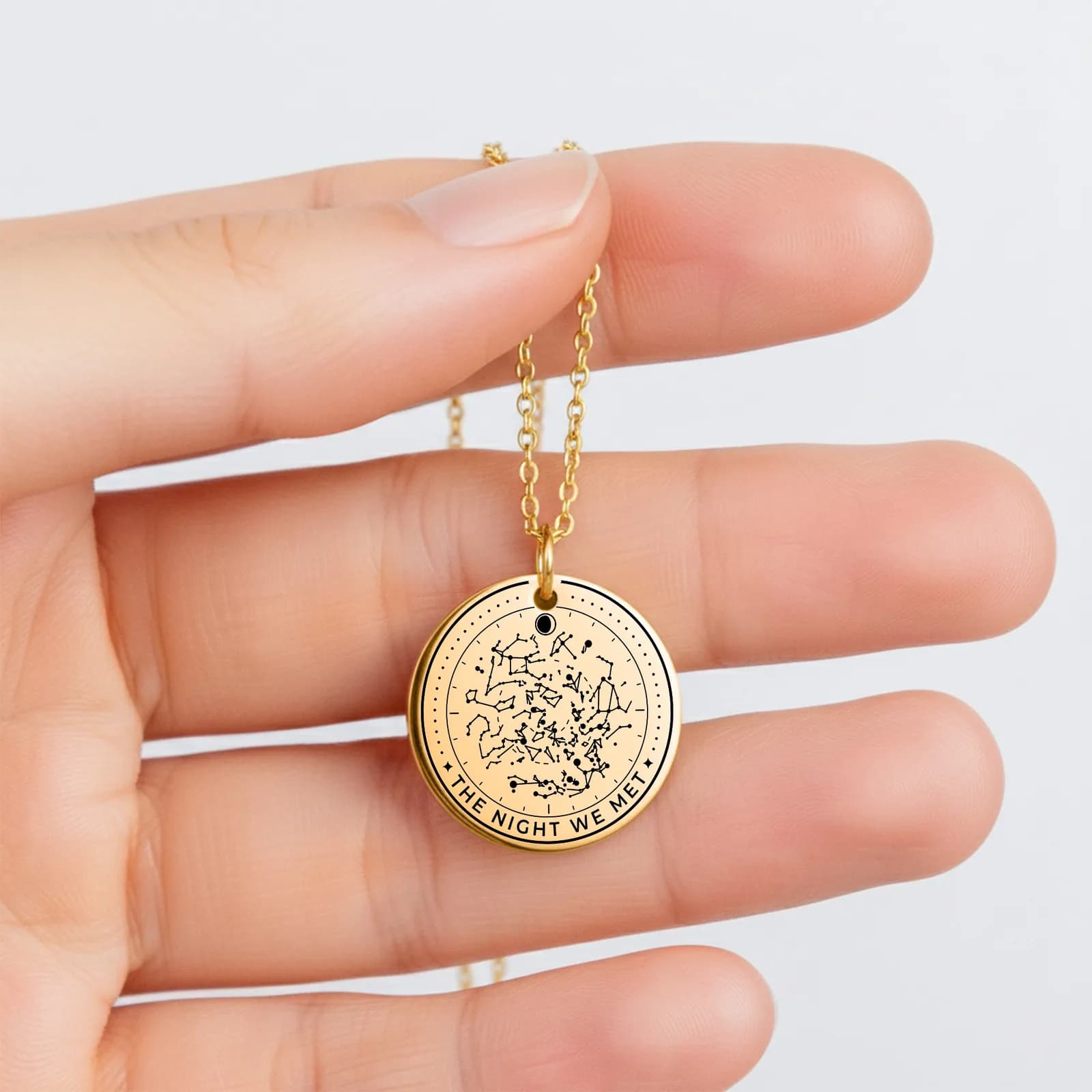 A gold star map coin necklace held between two fingers, engraved with constellations and the words The Night We Met